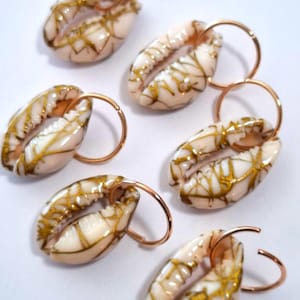 May include: Six cowrie shell earrings with gold-colored accents and copper-colored hoops. The shells are cream-colored with gold veining. The earrings are arranged on a white surface.