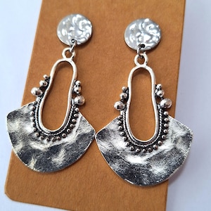 May include: A pair of silver-toned earrings with a hammered texture. Each earring features a circular top and a fan-shaped bottom with a decorative beaded edge. The earrings are displayed on a brown card.