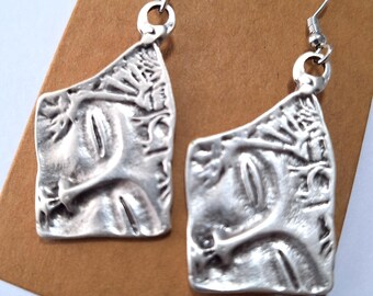 May include: A pair of silver earrings with a textured, abstract face design. The earrings are on a brown card.