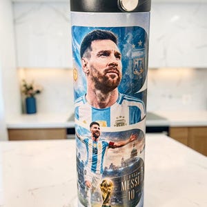 May include: A cylindrical water bottle with a black lid featuring a graphic print of Lionel Messi in an Argentina football shirt. The design includes the player's portrait, a full-body action shot, the World Cup trophy, and the text Messi 10.