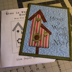 Puede incluir: Un tapiz de pared acolchado azul y verde con un diseño de casa de pájaros y el texto "Home is where you nest".