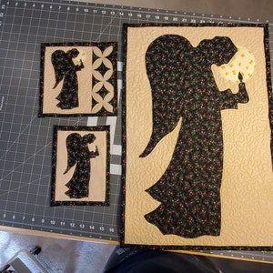 K&ouml;nnte beinhalten: Drei gequiltete Wandbeh&auml;nge mit Engel-Silhouetten. Der gr&ouml;&szlig;te Engel h&auml;lt eine Kerze und ist von einem beigefarbenen Hintergrund mit einem Blumenmuster umgeben. Die beiden kleineren Engel befinden sich auf beigefarbenen Hintergr&uuml;nden mit einem geometrischen Muster.