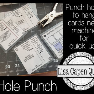 May include: A close-up of a hole punch tool on a cutting mat with several cards labelled with sewing machine instructions. The cards have instructions for different seam allowances and needle positions. The text on the cards reads "Move needle and align with centre of this line", "Line up edge of presser foot to right edge of this line", "Standard seam allowance used in quilting", "Test Seam Allowance", "Cut 3 squares exactly", "Seam = 1/4", "New needle position from machine needle", "1/4" Seam Allowance", and "Hole Punch". The text on the cards is in black and white. The cards are on a grey cutting mat with white markings. The hole punch is silver and has a black handle. The text "Punch holes to hang cards near machine for quick use" is in white text on a black background with a white circle with black dots around it. The text "Lisa Capen Quilts" is in white text on a black background inside the circle.