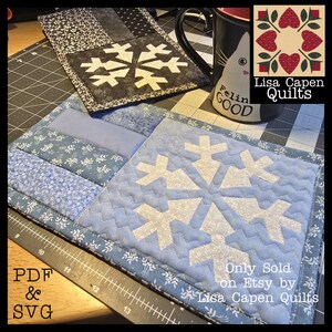 Peut inclure: Deux sets de table matelassés avec des motifs de flocons de neige. L'un est bleu clair avec un flocon de neige blanc, l'autre est noir et gris avec un flocon de neige blanc. Les sets de table sont sur un tapis de coupe. Le texte sur l'image indique "PDF & SVG" et "Only Sold on Etsy by Lisa Capen Quilts."