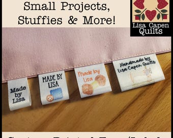 May include: Four different custom printed fabric tags with various designs. The tags are white with black text and feature images of sewing machines, yarn, and a sewing needle. The text on the tags reads "Made by Lisa", "Made by Lisa", "Handmade by Lisa Capen Quilts", and "MADE BY LISA". The tags are displayed on a pink fabric background with the text "For Quilts, Clothing, Small Projects, Stuffies & More!" and "Lisa Capen Quilts" above the tags. The text "Custom Printed Tags/Labels" is displayed below the tags.