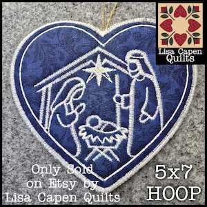 May include: A heart-shaped Christmas ornament with a blue fabric background and white embroidered nativity scene. The ornament has a white border and a gold string for hanging. The text "Lisa Capen Quilts" and "5x7 HOOP" are also visible.