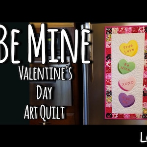 May include: A Valentine's Day art quilt with a pink, white, and red patchwork border. The center features four heart-shaped patches with the words "True Love", "Be Mine", "XOXO", and "Kiss Me" in pink lettering.