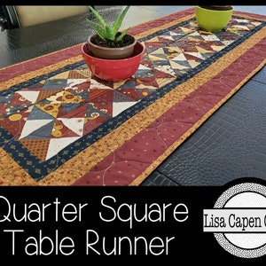 May include: A quilted table runner with a patchwork design featuring red, blue, and yellow fabrics. The runner has a burgundy border with a gold trim. The text "Quarter Square Table Runner" and "Lisa Capen Quilts" is visible in the image.