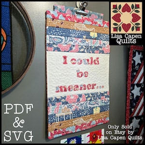 Könnte beinhalten: Ein gestecktes Klemmbrett mit dem Text "I could be meaner..." in roter Stickerei. Das Klemmbrett hat eine Mischung aus floralen und gemusterten Stoffen in Blau-, Rosa- und Brauntönen. Eine Metallklammer hält die Oberseite.