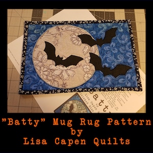 May include: A blue and white fabric mug rug with a moon and bats design. The text "Batty" Mug Rug Pattern by Lisa Capen Quilts" is printed below the mug rug.