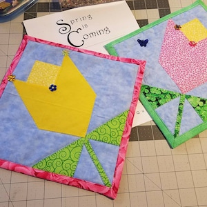 Pode incluir: Dois quadrados de tecido feitos à mão com um design de tulipa. Um quadrado é azul com uma tulipa amarela e folhas verdes. O outro quadrado é azul com uma tulipa rosa e folhas verdes. Ambos os quadrados têm uma borda rosa e verde.