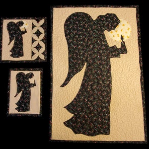 K&ouml;nnte beinhalten: Drei gequiltete Wandbeh&auml;nge mit der Silhouette eines Engels, der eine Kerze h&auml;lt. Der Engel ist schwarz mit einem Blumenmuster und der Hintergrund ist beige.