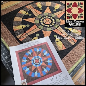 May include: A quilt pattern for a Mariner's Compass quilt titled "Show Me The Way". The quilt is 22 inches by 22 inches and features a starburst design with a variety of colours, including orange, red, blue, and black. The pattern is available for sale on Etsy.