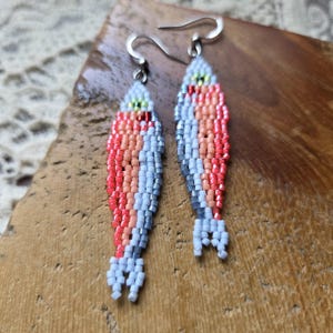May include: A pair of fish-shaped dangle earrings crafted from small glass beads in shades of coral, light pink, white, and grey. Each earring features a silver-toned metal hook and a detailed beaded pattern that mimics the scales and tail of a fish.