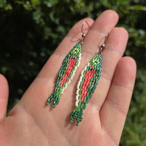 以下が含まれることがあります： ビーズで作られた魚の形をしたイヤリングのペア。イヤリングは、赤、緑、白のビーズのデザインが特徴です。魚の形をしたイヤリングには、銀色のイヤーフックが付いています。