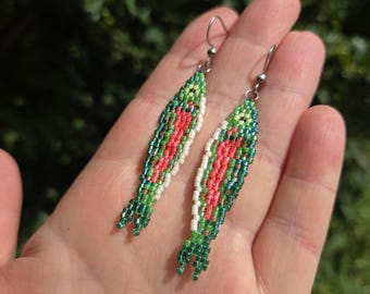 May include: A pair of beaded earrings in the shape of fish. The earrings feature a design with red, green, and white beads. The fish-shaped earrings have silver-colored ear wires.