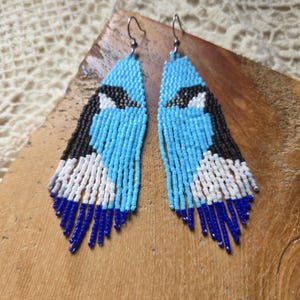 May include: Handmade beaded earrings featuring a blue jay design. The earrings are primarily light blue with black, white, and dark blue accents. The beaded fringe adds movement and detail to the unique, nature-inspired jewelry.