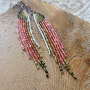 May include: A pair of beaded earrings with a fringe design. The earrings feature a combination of pink, white, yellow, green, and black beads. The earrings are displayed on a wooden surface.
