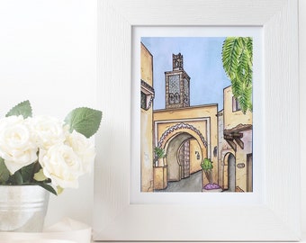 May include: A framed watercolor print of a Middle Eastern street scene. The artwork features buildings with arched doorways and a tall minaret. The color palette includes shades of yellow, beige, and blue. A bouquet of white roses in a silver bucket is in the foreground.