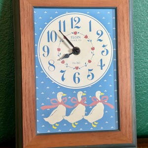 May include: Rectangular wooden-framed clock with a light blue face featuring a white clock dial with black hands and blue numerals. The clock face has a floral design and three white ducks with pink bows.