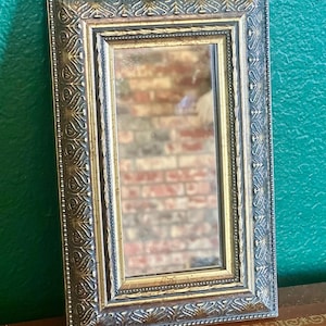 May include: A rectangular mirror with an ornate, gold-colored frame. The frame features detailed embossed patterns and a smaller, gold-colored inner frame. The mirror reflects a blurred image of a brick wall.