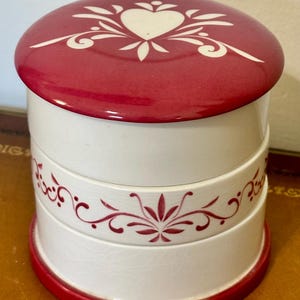 May include: A stacked, round ceramic container with a red lid and base. The lid features a white heart and floral design. The white body has red floral accents. The container is likely for storage or display.