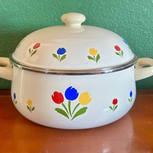 May include: White enamelware pot with lid and handles. The pot is decorated with colorful tulip flower designs in red, blue, and yellow. The pot has a silver rim and a cream-colored knob and handles.