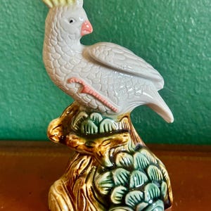 May include: A ceramic figurine of a white cockatoo with a yellow crest, perched on a tree stump. The bird has a pink beak and feet. The tree stump is painted with brown, green, and blue hues.