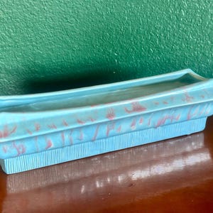 May include: A rectangular, turquoise ceramic planter with a textured base and a flared rim. The planter features a speckled pink pattern and is designed for displaying plants. The planter is a decorative item for home decor.