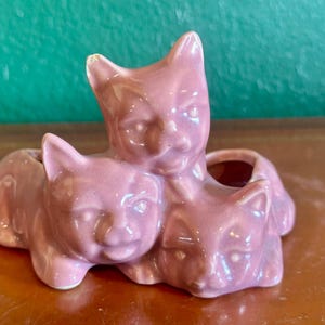 May include: A pink ceramic cat figurine with three cat figures. The cats are in a playful arrangement, with one cat standing and two cats lying down. The figurine is a decorative item.
