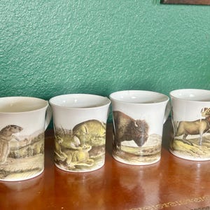 May include: Four white ceramic mugs with wildlife illustrations. Each mug features a different animal: a prairie dog, bison, deer, and ram. The mugs have a curved handle and a landscape scene. The mugs are displayed on a brown surface.