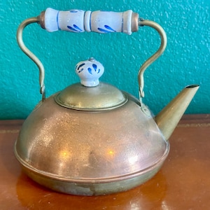 May include: A vintage copper teapot with a brass spout and handle. The handle features a white ceramic grip with blue floral designs. The lid is topped with a matching ceramic knob. The teapot is displayed on a brown surface against a teal background.