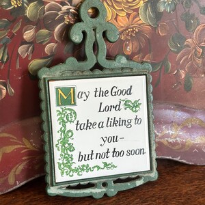 May include: Green metal pot holder with a white tile that reads "May the Good Lord take a liking to you - but not too soon."