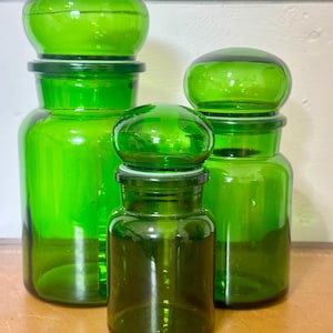 May include: Three emerald green glass apothecary jars of varying heights, each featuring a cylindrical body and a matching rounded, dome-shaped glass lid. The jars are transparent, smooth, and possess a uniform vibrant green color throughout.