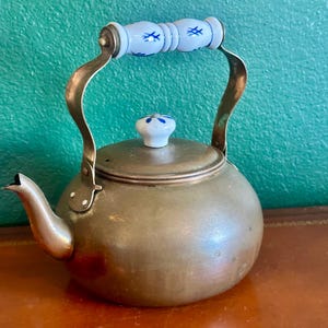 May include: A vintage brass teapot with a rounded body, a curved spout, and a hinged lid. The handle and lid knob are white ceramic with blue floral designs. The teapot sits on a brown surface against a teal green background.