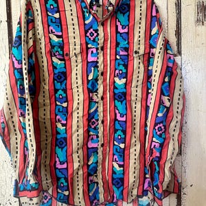 May include: A button-down shirt with a colorful geometric pattern. The shirt features vertical stripes in red, tan, and black, with blue, pink, and turquoise floral and geometric shapes. The shirt has two chest pockets and a collar.