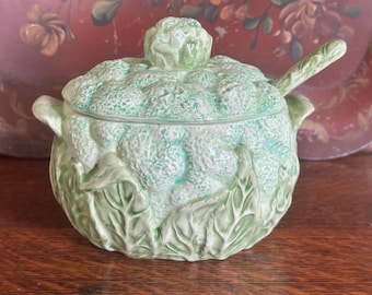 May include: A light green ceramic serving dish with a lid and a spoon. The dish is shaped like a head of cabbage, with detailed leaf and textured designs. The spoon rests on the side of the dish. A decorative kitchenware item.