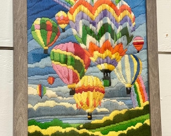 May include: A framed needlepoint artwork depicting a vibrant scene of hot air balloons floating in a blue sky with a rainbow. The balloons are various colors, including pink, green, yellow, and orange. The frame is a neutral gray.