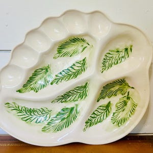 May include: Off-white ceramic serving platter with a leaf design. The platter has a scalloped edge and several compartments. Green leaf patterns are painted on the surface, adding a natural touch to the serving dish. Ideal for serving appetizers or snacks.