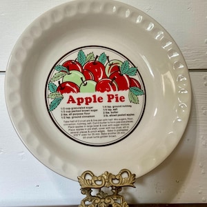 May include: A round, off-white ceramic pie dish with a scalloped rim. The center features a graphic of red apples with green leaves, the text "Apple Pie," and a printed recipe including ingredients like granulated sugar and ground cinnamon.