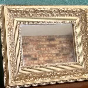 May include: A rectangular mirror featuring an ornate gold-colored frame with intricate embossed floral and scrollwork patterns. The frame has a multi-layered border design with a beaded inner edge and a wide, decorative outer molding.