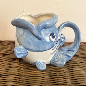 May include: A blue and white ceramic creamer shaped like a fish with a handle. The fish has a blue body with white accents and a blue tail fin. The creamer has a white interior and a blue rim.