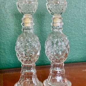May include: Two clear glass candle holders with a diamond pattern. The candle holders have a pineapple shape and a flared base. The top of the candle holders have a small cup shape to hold the candle. The candle holders are approximately 8 inches tall.