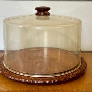 May include: A vintage cake stand with a clear glass dome and a wooden base. The base is round with a decorative, carved edge. The dome has a dark brown knob on top. The stand is set on a brown surface.