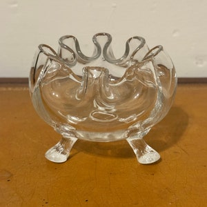May include: A clear glass bowl with a ruffled rim and three legs. The bowl is perfect for holding small items, such as jewelry or trinkets.