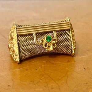 May include: A gold-tone metal miniature handbag brooch featuring a textured mesh body, a rigid top handle, and a decorative floral-shaped clasp centered with a small, round, vibrant green gemstone. The side panels display an ornate, raised relief pattern.