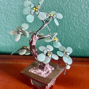 May include: A miniature decorative bonsai tree with light green jade-like leaves and yellow accents. The tree is set in a square, gold-toned metal pot with a textured base. The trunk is brown and the branches are made of wire.