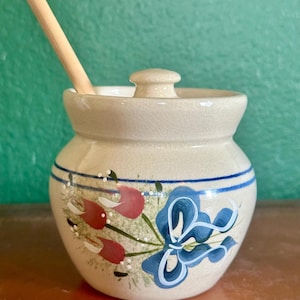 May include: A cream-colored ceramic jar with a lid and a wooden spoon. The jar is decorated with blue and red floral designs and two blue stripes. The jar is likely used for storing condiments or small food items.