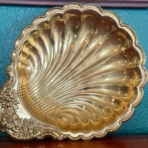 May include: A polished gold-tone metal dish shaped like a large scallop shell. The interior features deep, radiating fluted grooves, while the base area is decorated with intricate, raised floral and scrollwork relief detailing along the edge.
