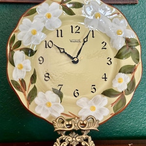 May include: A round wall clock with a pale yellow face, black numerals, and black hands. The clock features a hand-painted border of white flowers with yellow centers and green leaves. The brand name Verichron is printed in black on the clock face.